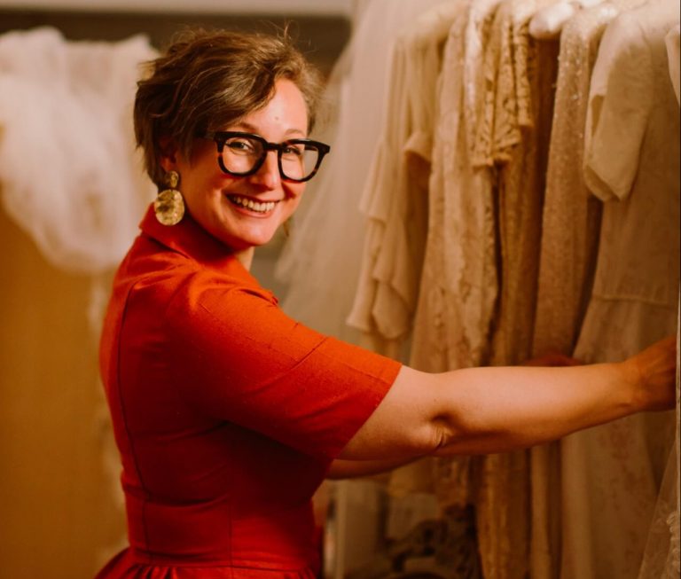 Kate Ashwell smiling, browsing a rail of vintage wedding dresses at Ashwell & Co