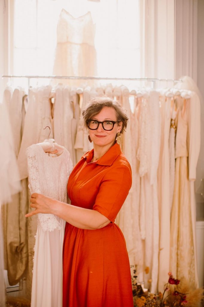 Kate Ashwell holding a vintage wedding dress, in front of a rail of vintage bridal dresses